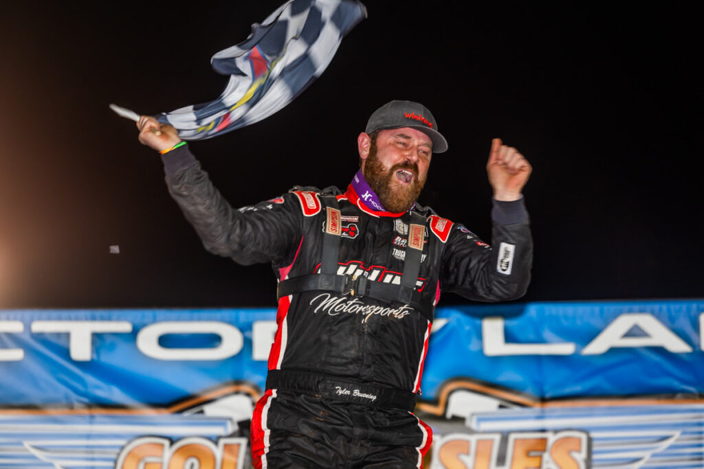 Tyler Bruening Achieves First Career Lucas Oil Victory waving the checkered flag with a happy expression on his face